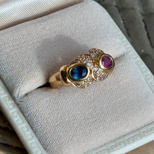 May include: A gold ring with a blue oval gemstone and a pink oval gemstone, both surrounded by small clear gemstones. The ring is set in a white velvet jewelry box.