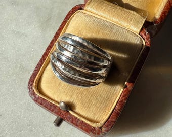 May include: A silver ring with a textured, layered design rests inside a vintage brown leather jewelry box with a gold satin interior. The ring's surface reflects light, highlighting its unique texture. The box is open, showcasing the ring.