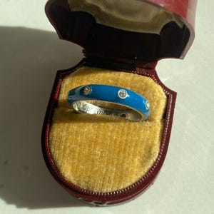 May include: A silver ring with a bright blue enamel band, set with three small diamonds. The ring is presented in a red jewellery box with a yellow velvet lining. The ring is a close-up shot.