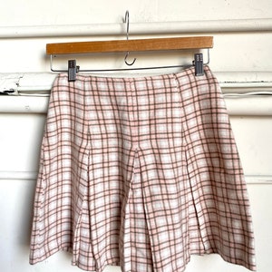 May include: A pink and brown plaid skirt with pleats. The skirt is hanging on a wooden hanger.