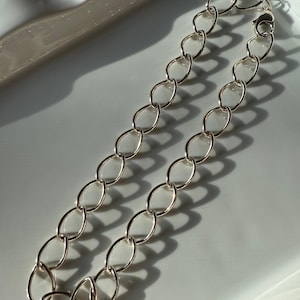May include: A silver-colored chain necklace with oval links. The necklace is displayed on a white surface, with shadows adding depth. The chain is a simple, elegant piece of jewelry.