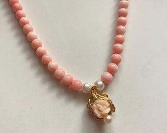 May include: A delicate pink coral bead necklace with a gold pendant. The pendant features a small pink rose and two white pearls. The necklace is a soft pink color and is displayed on a white surface.