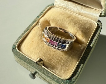May include: A silver ring with a row of colorful gemstones, including yellow, orange, pink, and blue, set in a band with a diamond-studded border. The ring is displayed in a vintage green jewelry box with a cream-colored interior.