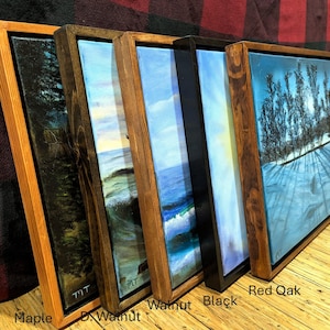 May include: Five rectangular canvas paintings in wooden floating frames, labeled Maple, D. Walnut, Walnut, Black, and Red Oak. The artworks depict various landscapes including a dark forest, ocean waves, and a snowy scene with trees and long shadows.