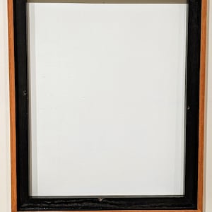May include: Square wooden picture frame with a black inner frame and a natural wood outer frame. The frame is empty, with a white interior. The frame is designed to hold a photograph or artwork.