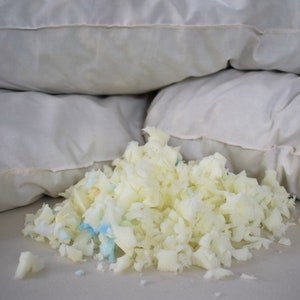 May include: A pile of white and blue shredded memory foam filling for a pillow. The foam is scattered on a white surface in front of three white pillows.