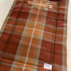 May include: A brown and green plaid dog bed with the name "Sherman" embroidered on it in gold thread. A paw print is also embroidered next to the name.