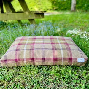 May include: A square pet bed with a plaid pattern in beige, claret, and white. The bed is placed on green grass. A small white tag with text is visible on the lower right corner. The bed is designed for pets.