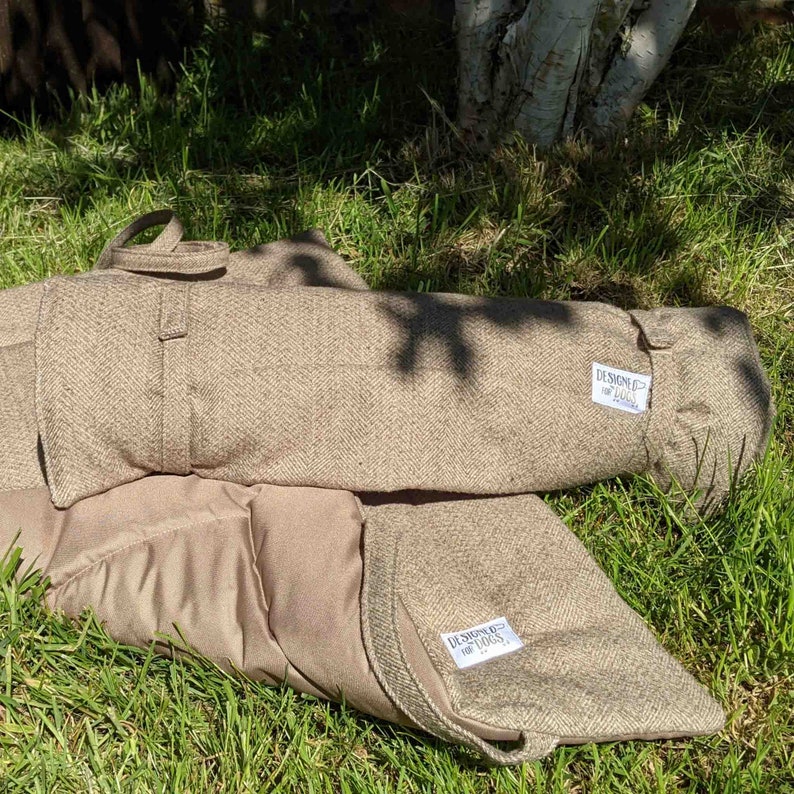 Designed for dogs picnic pad / travel mat in brown stone herringbone tweed, demonstrated rolled up and laid out on the grass. matching straps and handles, waterproof backing and lined with wadding for comfort.