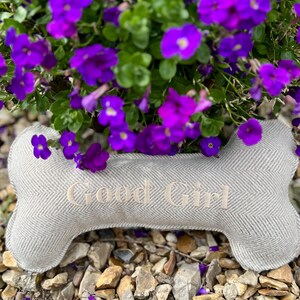May include: A gray and white herringbone dog bone pillow with the text "Good Girl" embroidered on it. The pillow is sitting on a bed of small gray rocks and is surrounded by purple flowers.