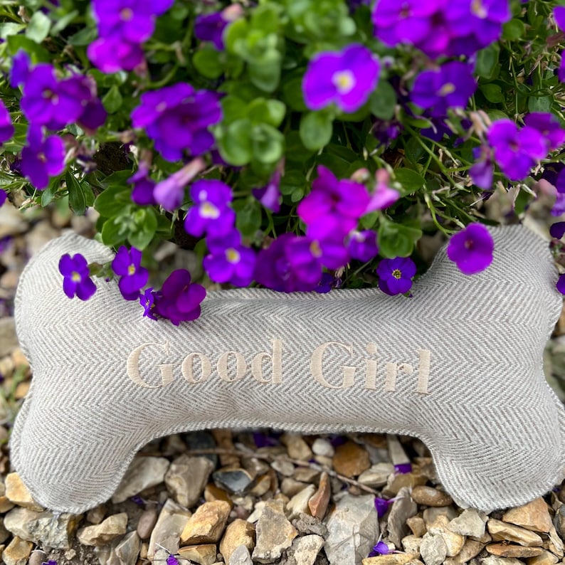 May include: A gray and white herringbone dog bone pillow with the text "Good Girl" embroidered on it. The pillow is sitting on a bed of small gray rocks and is surrounded by purple flowers.