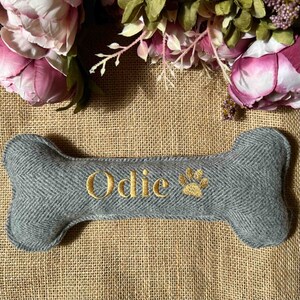 May include: Gray bone-shaped dog toy with the name "Odie" and a paw print embroidered in gold thread.