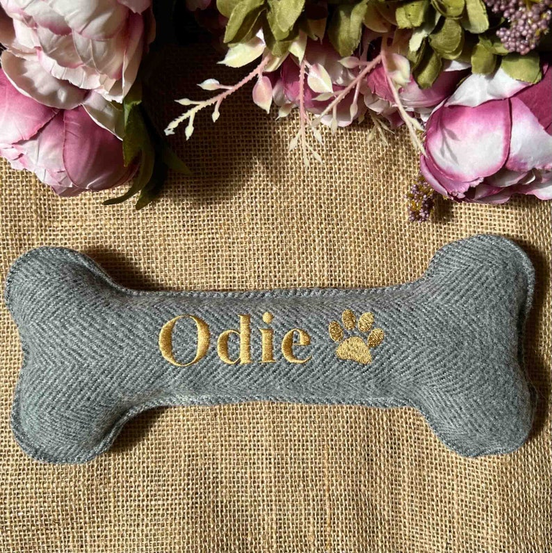 May include: Gray bone-shaped dog toy with the name "Odie" and a paw print embroidered in gold thread.