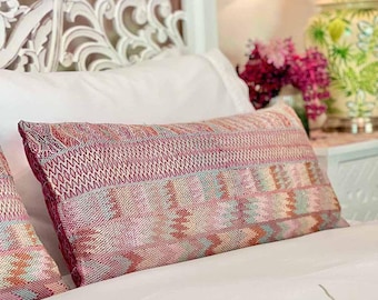 May include: A pink and white patterned throw pillow with a woven texture. The pillow is on a white bed with a white headboard.