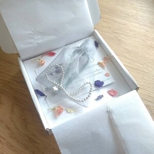 May include: A silver chain bracelet with a star charm, packaged in a white box with a delicate blue and white tulle bag. The box is filled with dried flower petals in shades of pink, purple, and white.