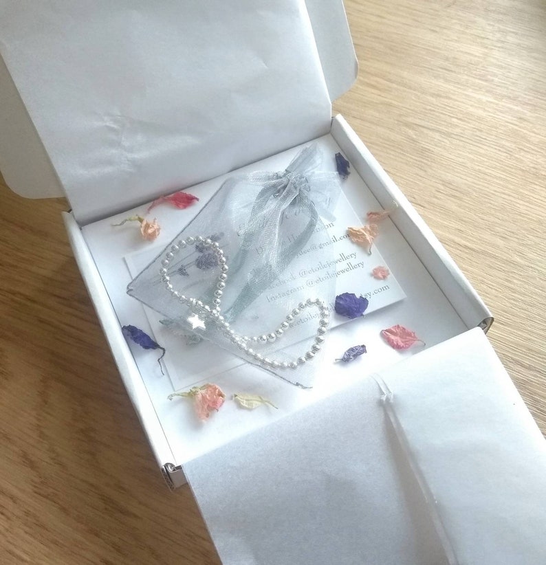 May include: A silver chain bracelet with a star charm, packaged in a white box with a delicate blue and white tulle bag. The box is filled with dried flower petals in shades of pink, purple, and white.