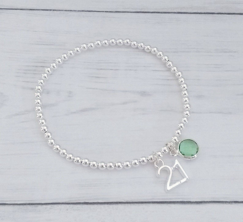 May include: A silver beaded bracelet with a silver charm that says "21" and a green gemstone charm.