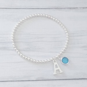 May include: A silver beaded bracelet with a charm featuring the letter A and a light blue gemstone.