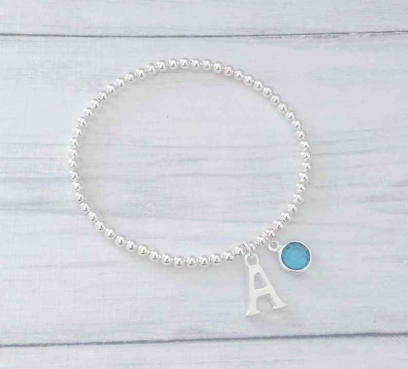 May include: A silver beaded bracelet with a charm featuring the letter A and a light blue gemstone.
