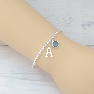 May include: A silver beaded bracelet with a charm featuring the letter 'A' and a blue gemstone.