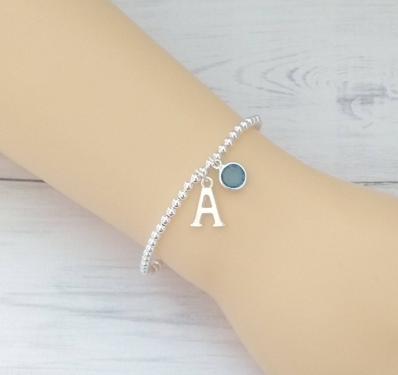 May include: A silver beaded bracelet with a charm featuring the letter 'A' and a blue gemstone.