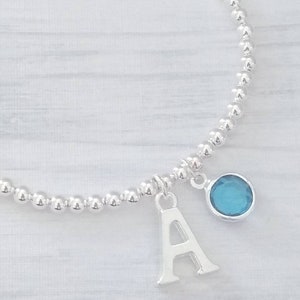 May include: A silver beaded bracelet with a charm featuring the letter A and a blue gemstone.