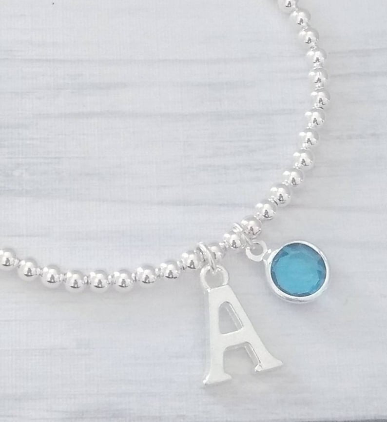 May include: A silver beaded bracelet with a charm featuring the letter A and a blue gemstone.