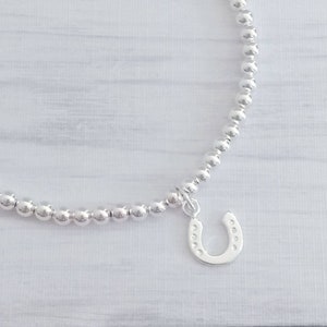 May include: A silver bracelet with a horseshoe charm. The bracelet is made of small, round silver beads. A horseshoe pendant hangs from the bracelet. The horseshoe charm is detailed and shiny, reflecting light.