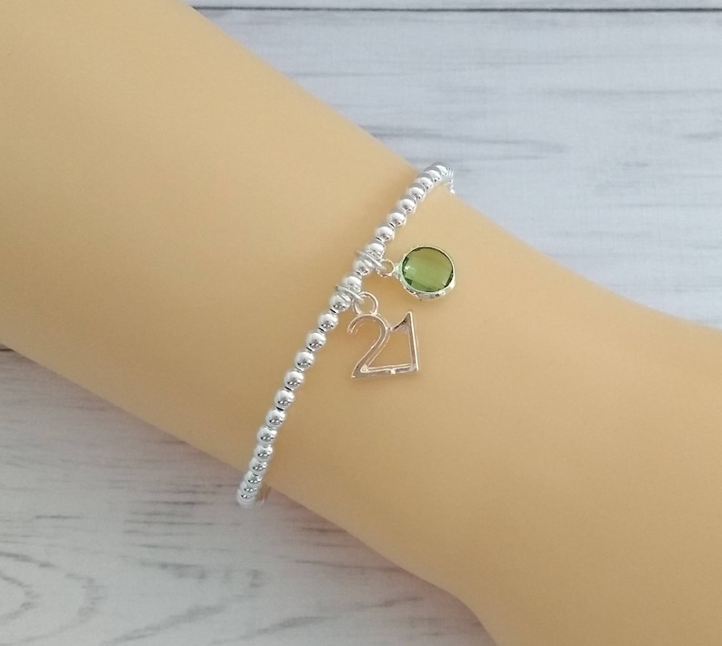 May include: A silver beaded bracelet with a green gemstone charm and a silver charm with the number 21.