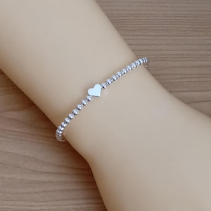 May include: A silver beaded bracelet featuring a small heart charm. The bracelet is composed of small, round beads. The heart charm is positioned centrally on the bracelet.