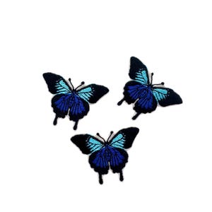 May include: Three embroidered butterfly patches featuring a black outer border with vibrant blue and light blue thread detailing on the wings. Each butterfly has two thin antennae and a symmetrical wing pattern with distinct dark and bright blue sections.