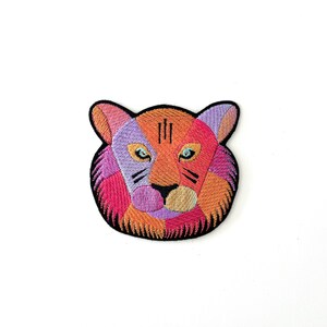 May include: A colourful embroidered patch depicting a tiger's face. The tiger has orange, pink, purple and blue fur. The patch is ideal for adding a touch of whimsy to clothing, bags or other accessories.
