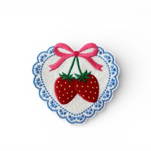 May include: Embroidered patch with two red strawberries, green stems, and a pink bow. The design is set on a white heart-shaped background with a blue scalloped border. Ideal for clothing or accessories.