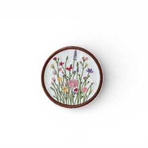 May include: A circular embroidered patch with a wildflower design. The patch has a white background and a brown border. The wildflowers include pink, yellow, purple, and red blooms with green stems.