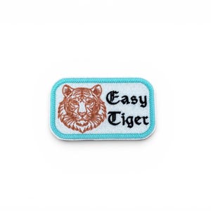 May include: Embroidered patch with a tiger face and the words "Easy Tiger" in black script. The patch has a white background, a light blue border, and a brown tiger design. The patch is rectangular with rounded corners.