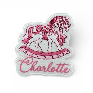 May include: An embroidered patch with a rocking horse design in pink and white. The horse has a pink mane, tail, and saddle. The name "Charlotte" is written in pink script below the horse.