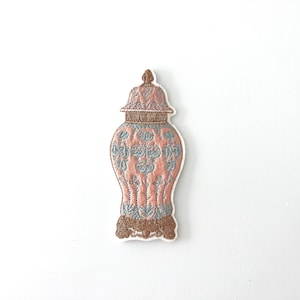 May include: Embroidered patch depicting a decorative ginger jar. The patch features a pink body with blue and brown embroidered details. The jar has a lid and sits on a brown base.