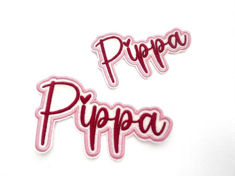 May include: Two pink and burgundy embroidered iron-on patches with the name "Pippa" in a cursive font. Each patch has a small heart in the middle of the name.