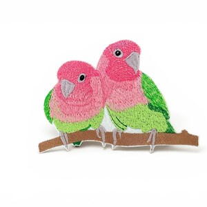 May include: An embroidered patch featuring two lovebirds perched on a brown branch. The birds have bright pink heads, light green wings, and grey beaks. The embroidery uses dense stitching to create a textured appearance of feathers on the birds.