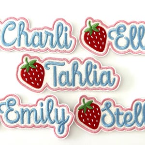 May include: Five embroidered iron-on patches with a strawberry design and a name in blue script. The names are Charli, Ella, Tahlia, Emily, and Stella.