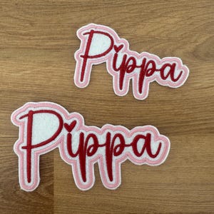 May include: Two pink and red felt patches with the name "Pippa" embroidered in red with a small heart in the middle of each patch.