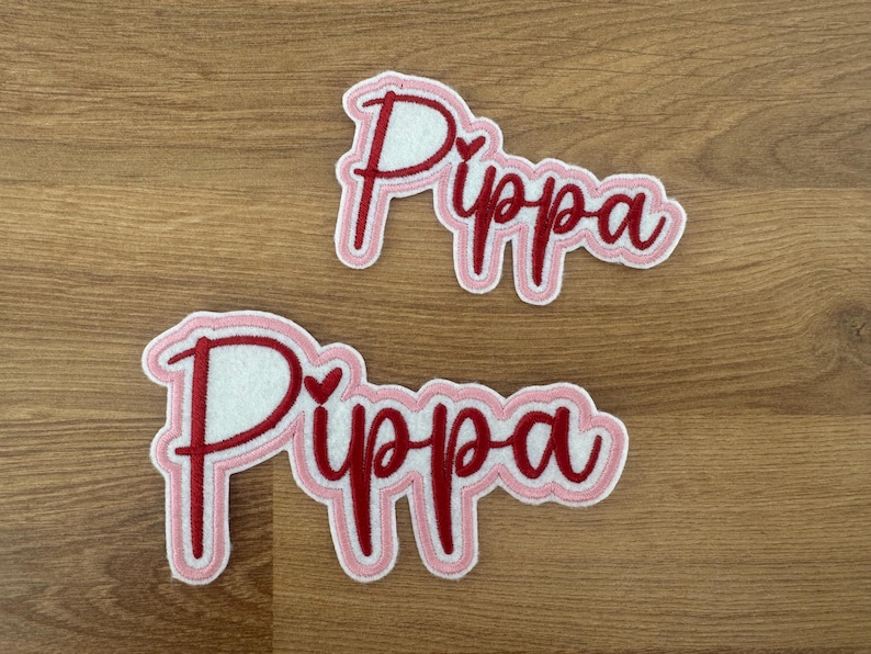 May include: Two pink and red felt patches with the name "Pippa" embroidered in red with a small heart in the middle of each patch.
