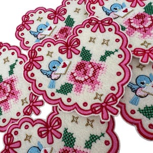 May include: Embroidered coasters with a scalloped edge. Each coaster features a pink rose, a blue bird carrying a letter, and pink bows. The coasters have a cream background with pink and green accents.