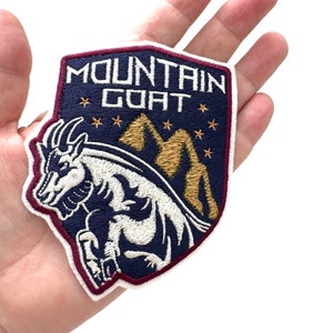 May include: An embroidered patch in navy blue with a white mountain goat and gold mountains and stars. The patch reads "Mountain Goat" in white lettering.