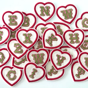 May include: A collection of felt heart-shaped patches with embroidered letters in green on a white background. The hearts have a red border.