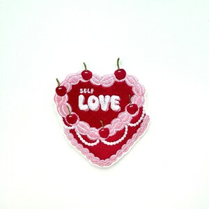 May include: Embroidered iron-on patch shaped like a heart-shaped cake with pink frosting and red cherries. The cake says "SELF LOVE" in white lettering.
