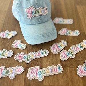 May include: A set of 12 colourful embroidered iron-on patches featuring the names of the zodiac signs. The patches are on a wooden surface with a blue denim baseball cap with a "Gemini" patch on the front.