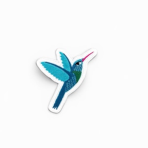 May include: An embroidered patch in the shape of a hummingbird. The bird features vibrant shades of blue and teal on its wings and body, with a small green patch on its chest. It has a thin, pointed beak in a bright pink colour and a white border.