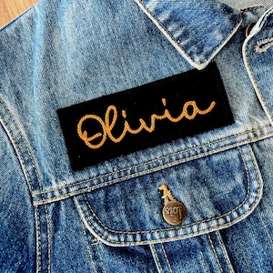May include: A black denim jacket with a black patch sewn on the front pocket. The patch has the name "Olivia" embroidered in gold thread.