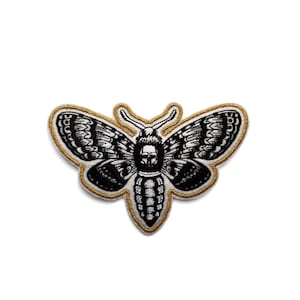 May include: Embroidered patch with a black and white moth design, showcasing detailed wing patterns. The moth is framed by a gold border. Ideal for customising clothing or accessories.